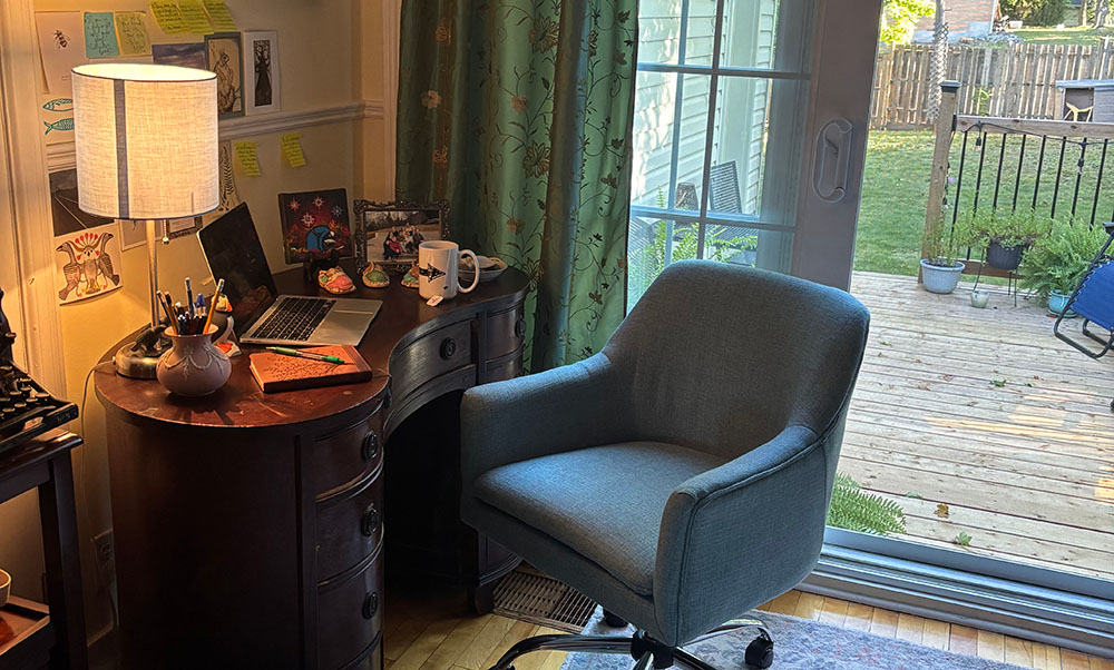 A grey office chair is positioned in front of an antique desk with a laptop and a cup on it. The desk is also adorned with a picture and a lamp. Through a sliding patio door the room opens into a large green yard. Some potted plants can be seen on the edge of the deck by the railing.