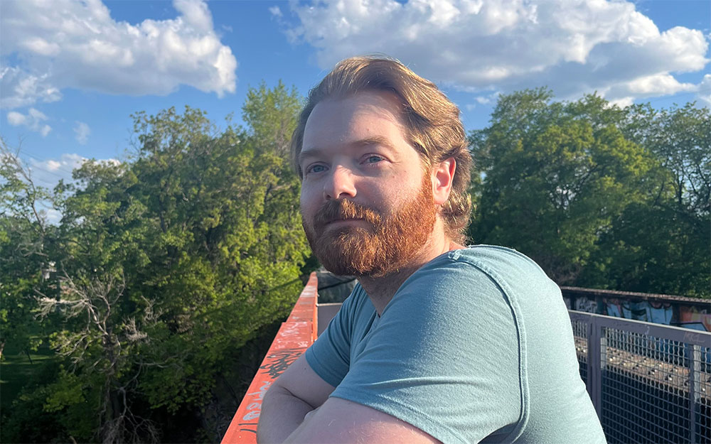 Jason Pchajek posing with a foot bridge and trees in the background.